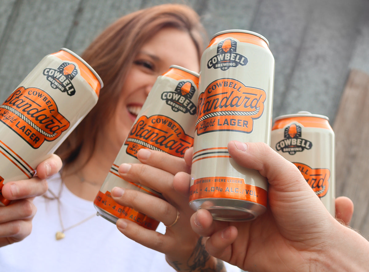 Four cans of Cowbell Standard Light Lager being held by people against a neutral background