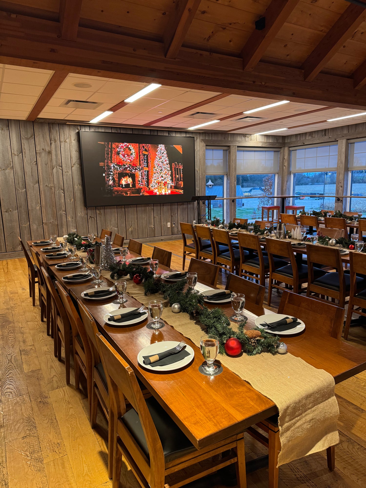 A large wooden dining room set up for a holiday gathering, with a long table covered in white plates, black napkins, and a burlap table runner decorated with green garlands and small ornaments. Multiple matching wooden dining tables and chairs are arranged in the room, which features wood-paneled walls, a wood ceiling with exposed beams, and large windows.